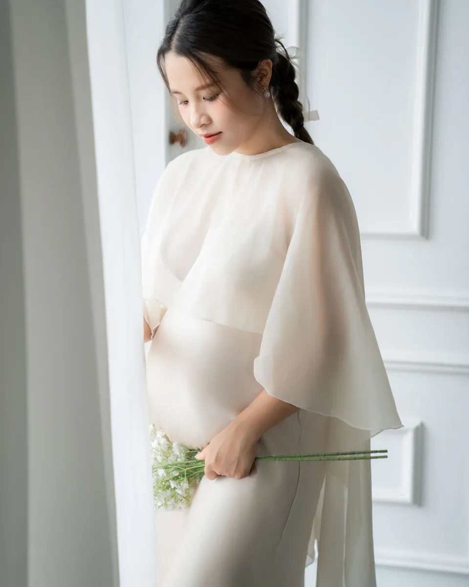 Future maman de profil près d’une fenêtre, vêtue d’une robe claire
